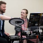 student using the drums, with a teacher helping out